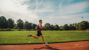 A runner strides along a red track beside a park field.