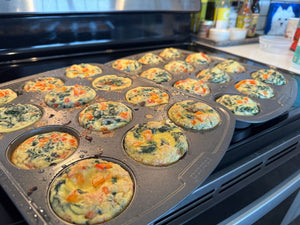 A dozen golden spinach and feta egg bites fresh out of the oven in a muffin tray, flecked with mushrooms, red bell pepper, and wilted spinach